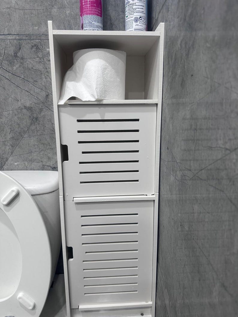 May include: A white, freestanding bathroom storage cabinet with a slatted door design. The cabinet has a shelf holding a roll of toilet paper and two bottles. The cabinet is next to a white toilet.