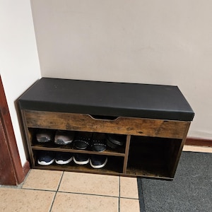 May include: A wooden shoe storage bench with a black cushioned top. The bench has two shelves and a compartment for shoe storage. Several pairs of shoes are visible on the shelves. The bench is made of brown wood.