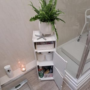 Op de afbeelding: Een witte, drielaagse opbergkast met een varen in een witte pot bovenop. De kast heeft een rieten mand, twee witte bakjes en een kleine open deur met schoonmaakmiddelen. Een zeester siert de bovenste plank.