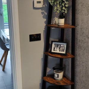 Puede incluir: Estantería esquinera de madera con cuatro estantes y soportes metálicos negros. Exhibe una foto enmarcada, una vela, una caja y plantas en macetas. La unidad se coloca contra una pared gris, ideal para objetos decorativos.