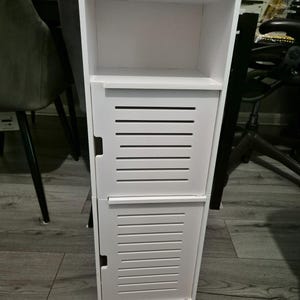 May include: A tall, white wooden storage cabinet with two slatted doors and an open shelf. The cabinet is designed for vertical storage and has a clean, modern aesthetic. It is suitable for bathroom or bedroom use.