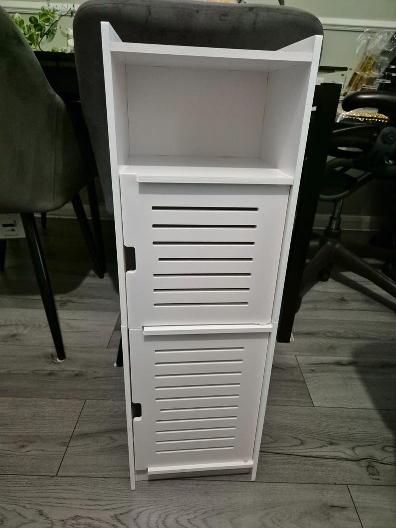May include: A tall, white wooden storage cabinet with two slatted doors and an open shelf. The cabinet is designed for vertical storage and has a clean, modern aesthetic. It is suitable for bathroom or bedroom use.