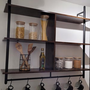 Può includere: Una mensola da cucina marrone scuro con supporti in metallo nero. I ripiani contengono barattoli di vetro con pasta, spezie e tè, utensili di legno, olio d'oliva e tazze nere appese sotto. L'unità è montata su una parete bianca.