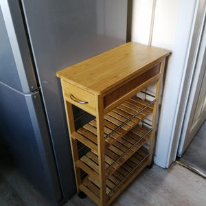 May include: A natural bamboo rolling storage cart with a drawer and three open shelves. The cart has a rectangular top and is supported by a wooden frame with metal bars. The cart is on small black wheels.