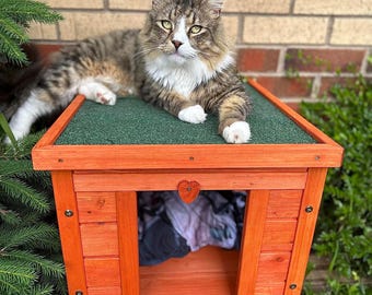 May include: A fluffy, multi-colored cat with white paws and chest rests on top of a small, orange wooden cat house. The cat house has a green roof and a heart-shaped cutout. The cat house is outdoors, surrounded by greenery and a brick wall.