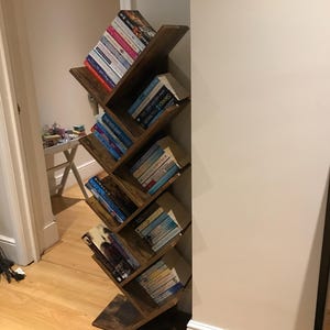 Op de afbeelding: Een donkerbruine zigzag-boekenkast gevuld met diverse boeken. De houten boekenkast staat tegen een witte muur en heeft meerdere planken in afwisselende hoeken. De boeken zijn van verschillende formaten en kleuren.
