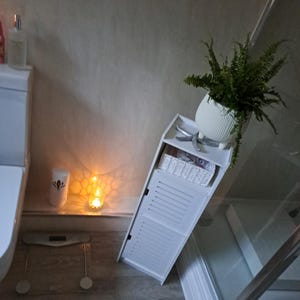 May include: A white bathroom cabinet with a slatted door, a white wicker basket, and a potted fern. A scale, a candle, and a bottle of lotion are also visible. The cabinet is angled.