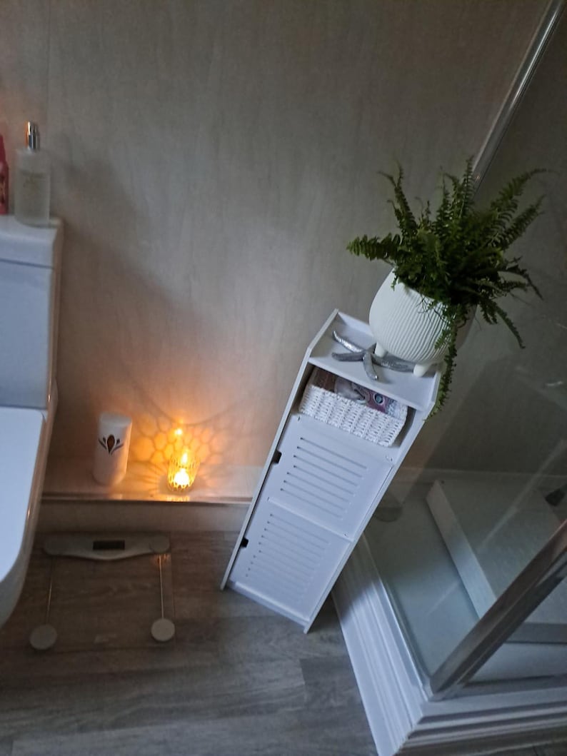 May include: A white bathroom cabinet with a slatted door, a white wicker basket, and a potted fern. A scale, a candle, and a bottle of lotion are also visible. The cabinet is angled.