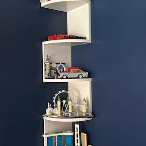 Op de afbeelding: Een witte hoekplank toont miniatuur gebouwmodellen en speelgoedauto's. De bovenste plank heeft een blauw en wit huismodel. Andere planken tonen een rode auto, een Londense scène en een stadsgezicht van Singapore.