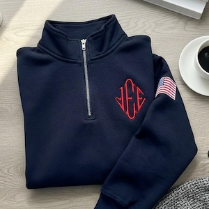 Könnte beinhalten: Ein marineblaues Sweatshirt mit einem Reißverschlusskragen und einem roten Monogramm auf der Brust. Auf dem Ärmel befindet sich ein amerikanischer Flaggenaufnäher. Eine Tasse Kaffee, ein Smartphone und eine Pflanze sind ebenfalls zu sehen.