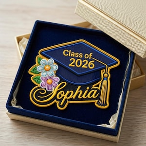 Può includere: Toppa ricamata con cappello di laurea in una confezione regalo. Il cappello blu navy presenta finiture dorate, il testo "Class of 2026" e il nome "Sophia". Accenti floreali in azzurro, viola e giallo circondano il cappello. La confezione regalo è beige.