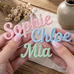Könnte beinhalten: Gestickte Namensaufnäher in Rosa, Blau und Grün. Die Namen „Sophie“, „Chloe“ und „Mia“ sind in Schreibschrift dargestellt. Die Aufnäher liegen auf einer Holzoberfläche, im Hintergrund eine kleine Vase und Trockenblumen.