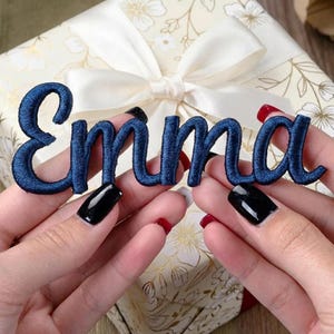 Puede incluir: Nombre bordado en azul marino "Emma" en una fuente cursiva. Las letras se sostienen sobre una caja de regalo con un lazo blanco. La caja de regalo tiene un estampado floral.