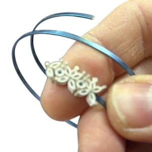 May include: A blue metal bracelet with a white floral design. The bracelet is held in a hand, highlighting the intricate details of the jewellery. The floral design is delicate and adds a touch of elegance to the bracelet.
