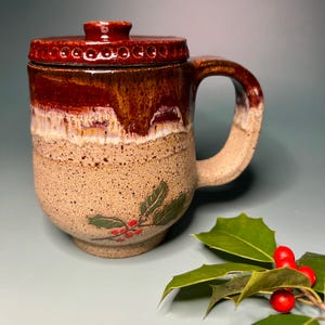 May include: A handmade ceramic mug with a lid, featuring a brown and tan glaze. The mug has a handle and is decorated with a holly leaf and berry design. The mug is approximately 4 inches tall.