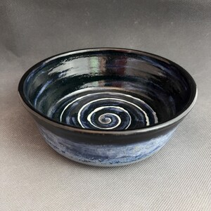 May include: A handmade ceramic bowl with a glossy black interior and a white spiral design at the bottom. The exterior has a gradient of blue and black. The bowl is suitable for serving food or as a decorative piece.