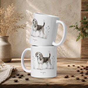 May include: Two white ceramic mugs stacked vertically, each featuring a detailed black and white illustration of a standing beagle dog surrounded by small floral sketches. Below each dog, the word beagle is printed with a small heart symbol.