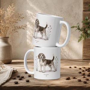 Puede incluir: Dos tazas de cerámica blanca apiladas verticalmente, cada una con una ilustración detallada de un perro beagle de pie entre flores silvestres estilizadas. Debajo de cada perro, la palabra beagle está impresa en pequeño con un corazón.
