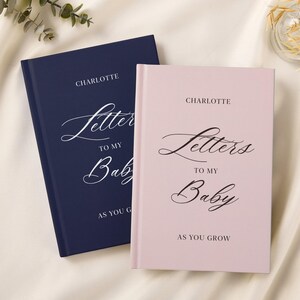 May include: Two baby journals, one navy blue and one blush pink. Each journal has the name "Charlotte" at the top, with the words "Letters To My Baby" and "As You Grow" in elegant script.