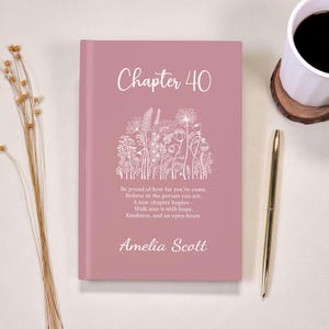 Peut inclure: Un livre rose poudré avec le titre "Chapter 40", des illustrations florales blanches et un texte inspirant. Un stylo doré et une tasse de café sont à proximité. Le nom "Amelia Scott" est également présent.