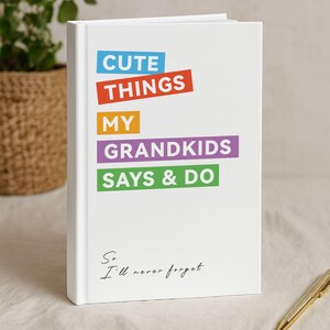 May include: A white hardcover journal with the text "CUTE THINGS MY GRANDKIDS SAYS & DO" in colorful block letters. The phrase "So I'll never forget" is written in cursive at the bottom. A gold pen rests on the surface.