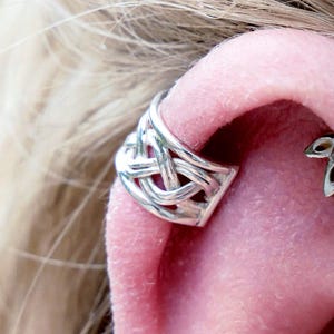 May include: Close-up of an ear with several pieces of jewellery. A silver ear cuff with a Celtic knot design is on the upper ear. A small, sparkling stud earring is visible in the lobe, and a floral-shaped earring is also present.