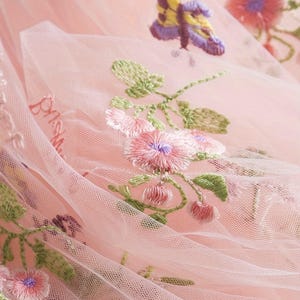 Peut inclure: Gros plan sur un tissu de tulle rose d&eacute;licat avec une broderie florale et papillon complexe. La broderie pr&eacute;sente des fleurs roses avec des centres violets, des feuilles vertes et un papillon aux ailes jaunes et violettes. Le tissu est l&eacute;g&egrave;rement translucide.