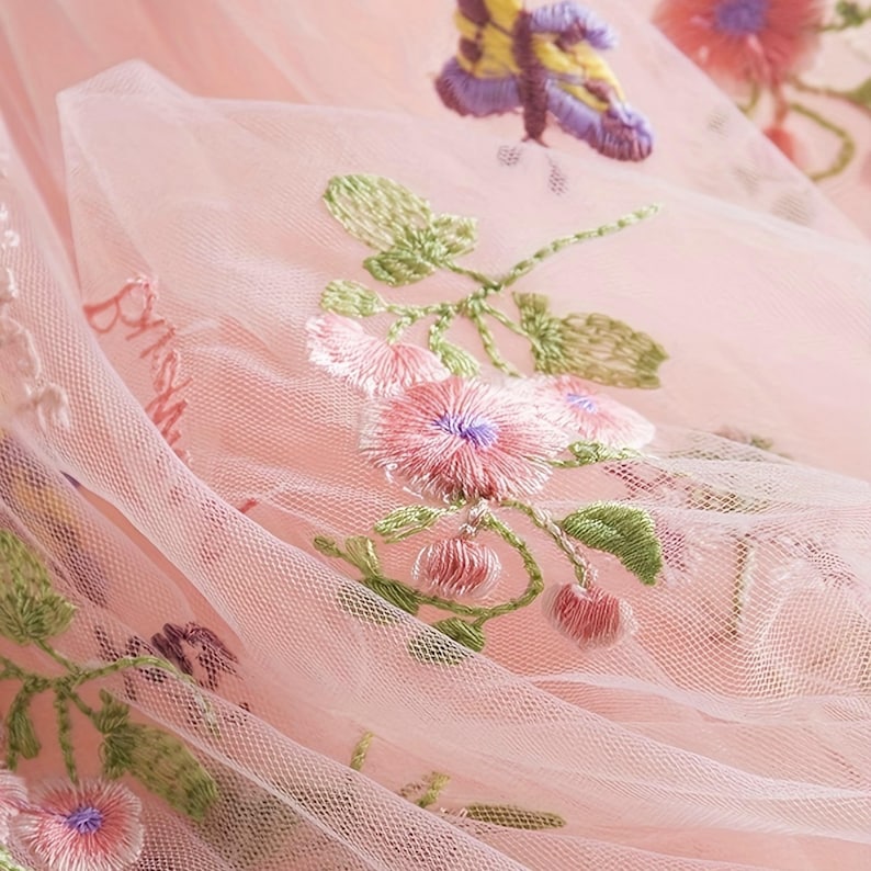 Peut inclure: Gros plan sur un tissu de tulle rose d&eacute;licat avec une broderie florale et papillon complexe. La broderie pr&eacute;sente des fleurs roses avec des centres violets, des feuilles vertes et un papillon aux ailes jaunes et violettes. Le tissu est l&eacute;g&egrave;rement translucide.
