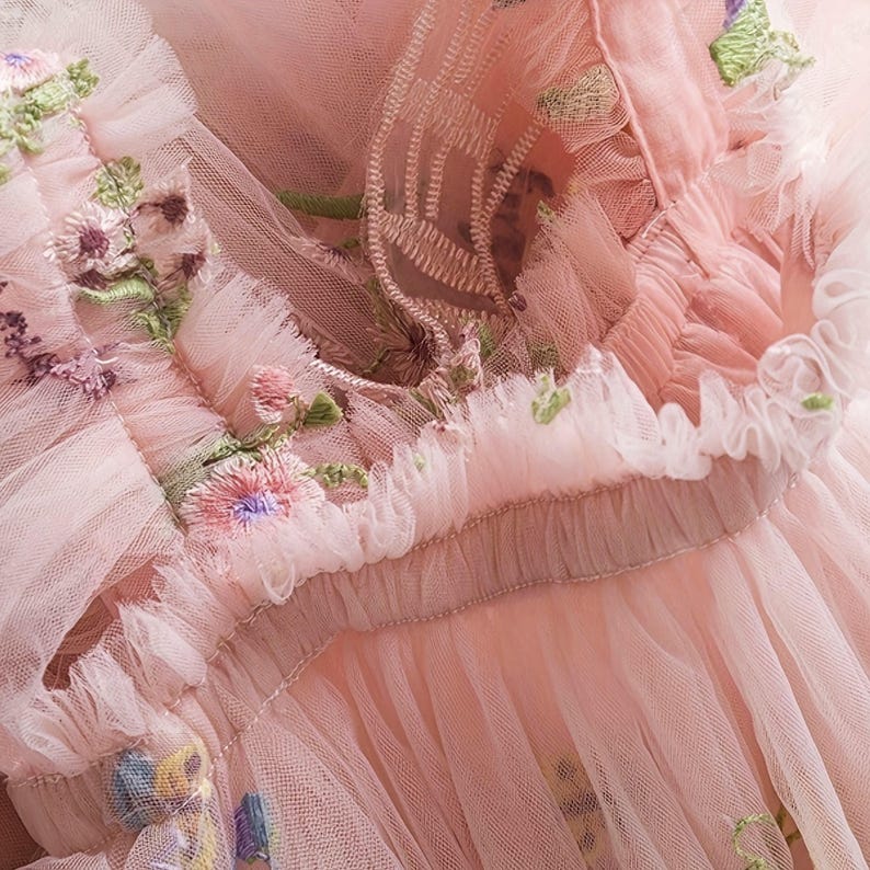 Peut inclure: Gros plan sur un tissu en tulle rose avec des broderies florales complexes. Le design d&eacute;licat pr&eacute;sente des couches de tissu fronc&eacute; et des coutures d&eacute;taill&eacute;es dans des tons de rose, de vert et de violet, cr&eacute;ant une esth&eacute;tique romantique et &eacute;th&eacute;r&eacute;e.