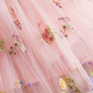 Peut inclure: Gros plan sur un tissu en tulle rose avec des fleurs et des papillons brod&eacute;s de diff&eacute;rentes couleurs. Le tissu a plusieurs couches, cr&eacute;ant un effet doux et fluide. La broderie comprend le mot "fairy" en cursive.