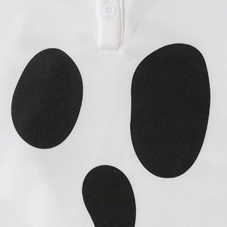 May include: A white polo shirt featuring a black graphic print of three large, irregular oval shapes arranged to resemble a face. The garment includes a white placket with a single visible white button at the top center of the neckline area.