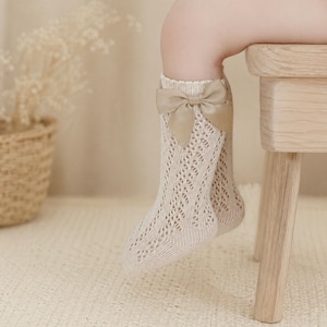 May include: A pair of beige knee-high socks with a delicate openwork design. Each sock features a large, light brown bow at the top. The socks are worn by a child, with their leg resting on a small wooden stool.