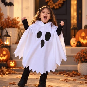 May include: A white hooded ghost poncho featuring a black printed face with two oval eyes and a teardrop-shaped mouth. The garment has a jagged hemline and is worn over a black long-sleeved shirt, black leggings, and black boots for a costume look.
