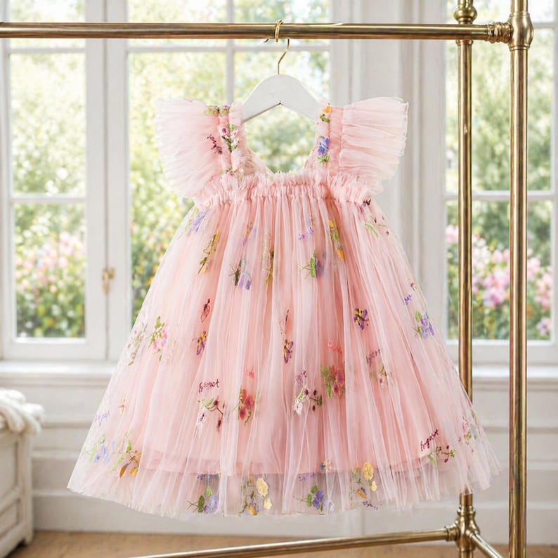 Peut inclure: Une robe en tulle rose clair avec broderies florales et manches &agrave; volants est suspendue &agrave; un cintre blanc. La robe pr&eacute;sente une encolure d&eacute;gag&eacute;e et une jupe &agrave; plusieurs &eacute;paisseurs, id&eacute;ale pour une occasion sp&eacute;ciale. La robe est expos&eacute;e sur un portant dor&eacute;.