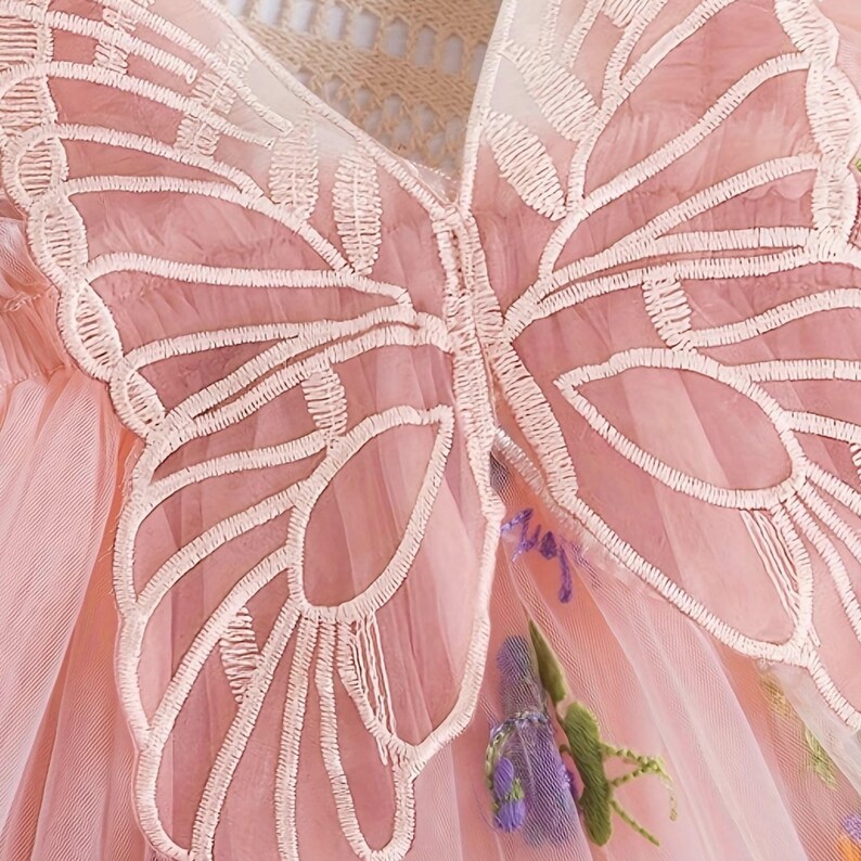 Peut inclure: Gros plan d'une robe rose avec un motif d'ailes de papillon. Les ailes sont soulign&eacute;es de broderies blanches et sont centr&eacute;es sur le corsage. La robe a un voile de tulle et est orn&eacute;e de broderies florales.