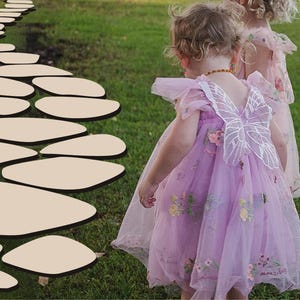 May include: Two young girls wearing fairy costumes walk on a path made of flat stones. The girl in the front is wearing a lavender dress with a white butterfly on the back. The girl in the back is wearing a pink dress with white polka dots.