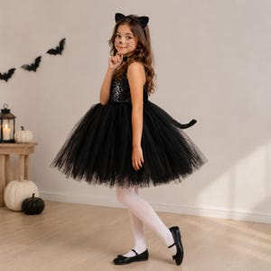 May include: A black cat costume featuring a sleeveless dress with a sequined bodice and a voluminous tulle skirt. The ensemble includes a black headband with cat ears and a matching tail attached to the back of the dress, paired with white tights.