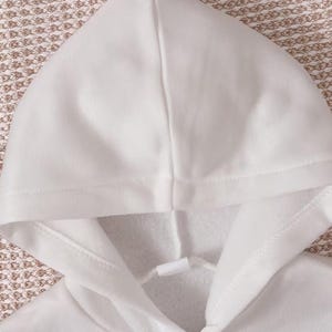 May include: A plain white hooded sweatshirt featuring a smooth exterior fabric and a soft, brushed interior lining. The hood includes a matching white drawstring with a plastic aglet at the center, providing a clean and simple aesthetic design.
