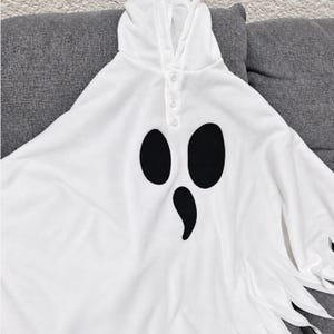May include: A white hooded poncho featuring a ghost face graphic with two large black oval eyes and a smaller black teardrop-shaped mouth. The garment includes a front placket with three white buttons and a hemline with jagged, pointed fabric edges.