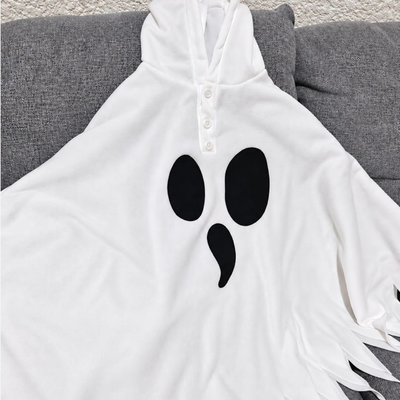 May include: A white hooded poncho featuring a ghost face graphic with two large black oval eyes and a smaller black teardrop-shaped mouth. The garment includes a front placket with three white buttons and a hemline with jagged, pointed fabric edges.