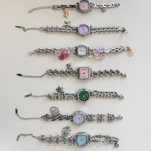 May include: A selection of eight silver-coloured bracelet watches with diverse face shapes and colours, including pink, green, and blue. Some watches have decorative charms such as stars and flowers. The watches are displayed on a white surface.