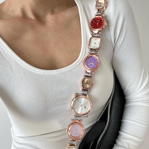 Könnte beinhalten: Ein einzigartiger Schulterriemen aus mehreren roségoldenen Uhren mit verschiedenen farbigen Zifferblättern, darunter Rot, Lila und Silber. Der Riemen ist an einer schwarzen Tasche mit einem silbernen Reißverschluss befestigt.