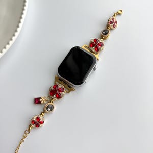 May include: Gold-toned Apple Watch band with red and clear jewel accents. The band features flower-shaped embellishments with red jewels, and a chain extension. The watch face is black.