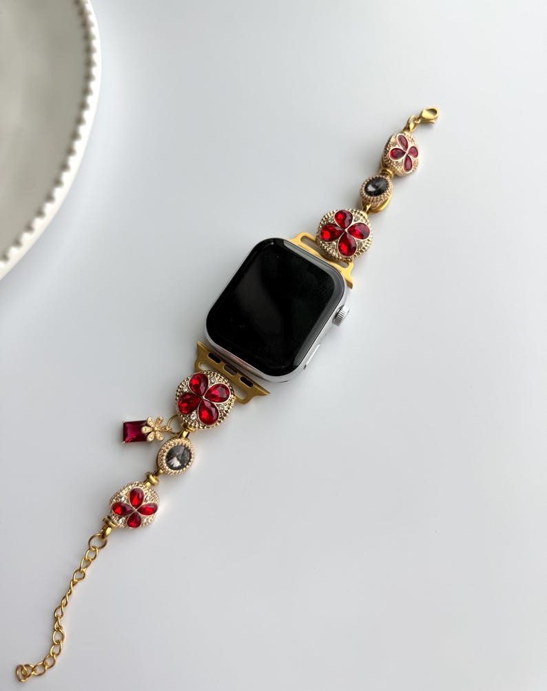 May include: Gold-toned Apple Watch band with red and clear jewel accents. The band features flower-shaped embellishments with red jewels, and a chain extension. The watch face is black.