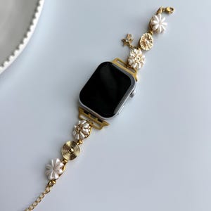 May include: A gold-toned Apple Watch with a decorative band. The band features white flower and pearl accents, along with a gold chain. The watch face is black. The band is connected to the watch with gold-colored connectors.
