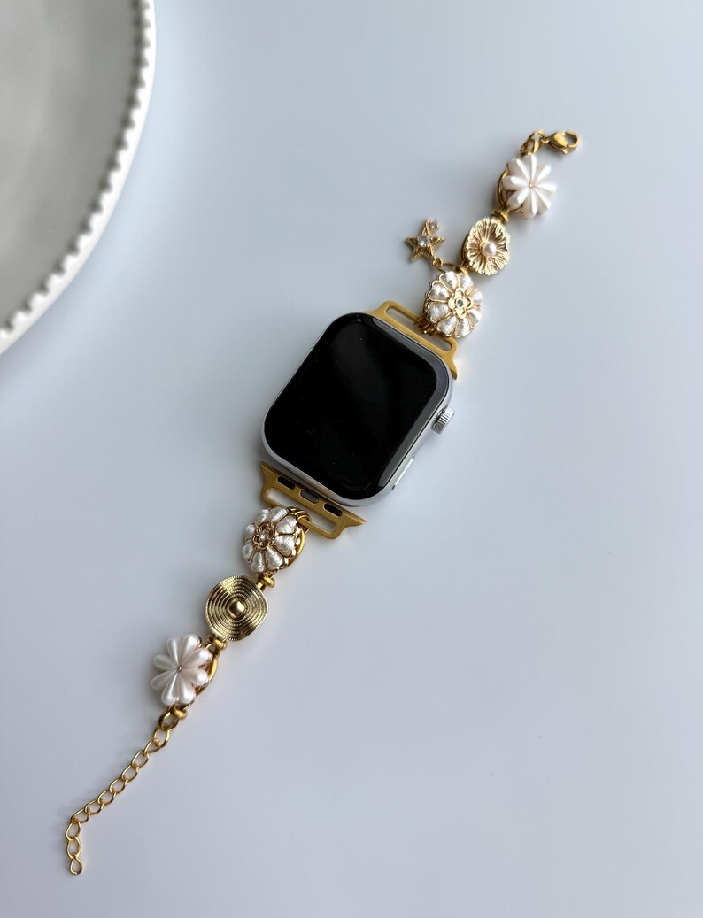 May include: A gold-toned Apple Watch with a decorative band. The band features white flower and pearl accents, along with a gold chain. The watch face is black. The band is connected to the watch with gold-colored connectors.