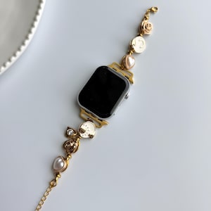 May include: Gold-toned Apple Watch band with decorative charms. The band features a chain, pearls, and ornate embellishments. The watch face is black, and the band is set against a white background.