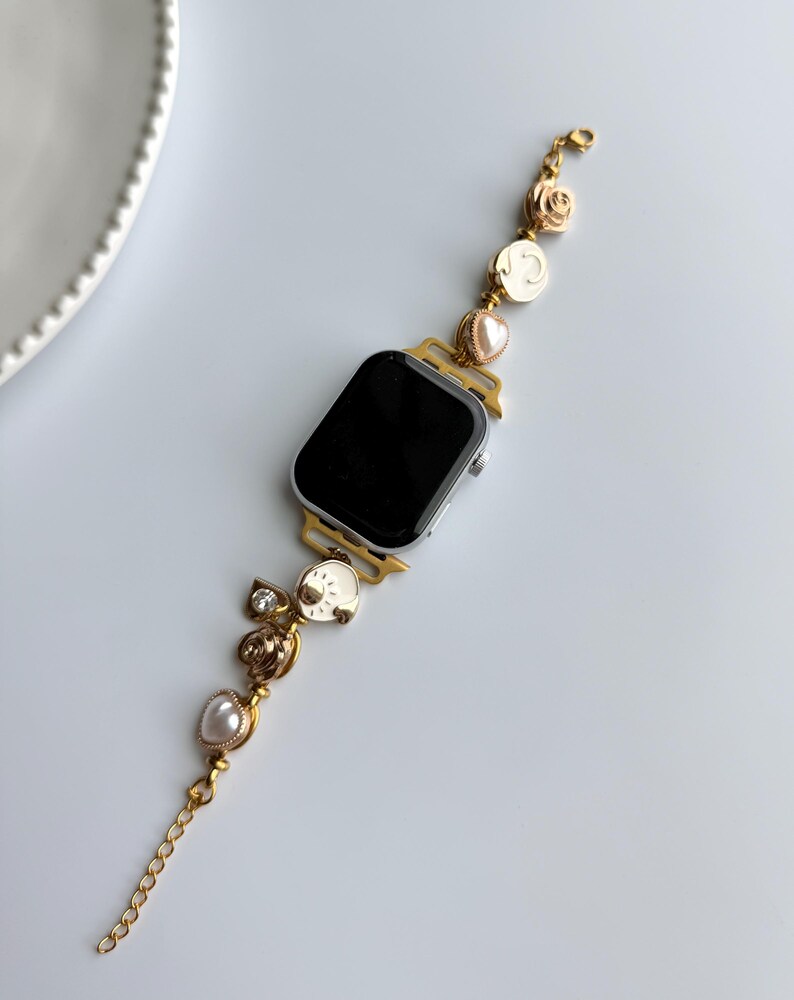 May include: Gold-toned Apple Watch band with decorative charms. The band features a chain, pearls, and ornate embellishments. The watch face is black, and the band is set against a white background.