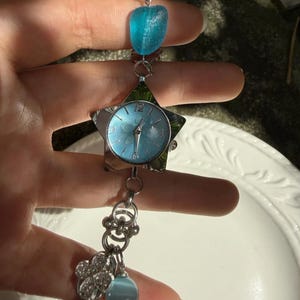 May include: A silver-toned watch with a star-shaped face and a light blue dial. The watch features a blue stone and a cluster of charms. The watch is held up by a hand.