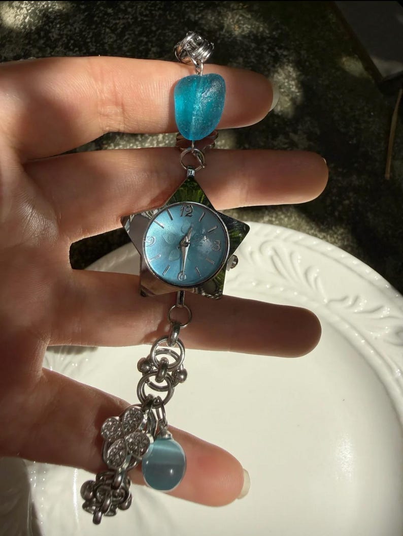 May include: A silver-toned watch with a star-shaped face and a light blue dial. The watch features a blue stone and a cluster of charms. The watch is held up by a hand.